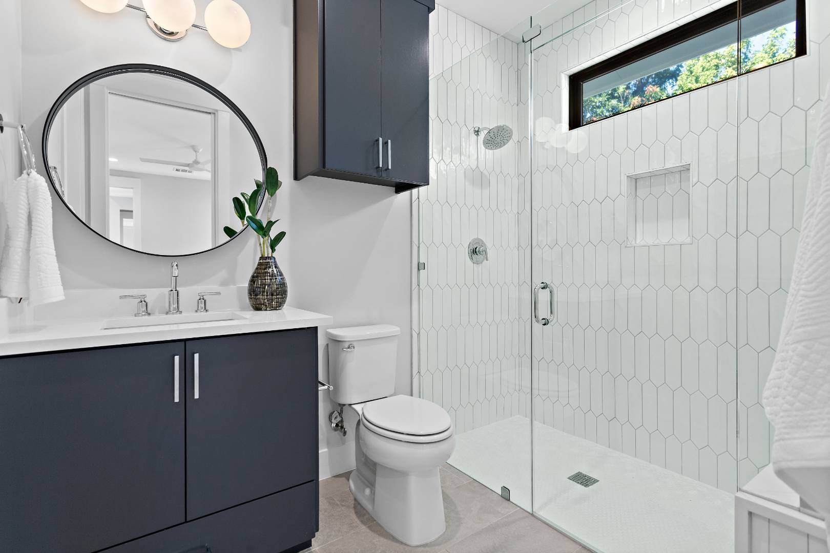 Modern bathroom with a sleek design: Features dark cabinetry, a round mirror, and a glass-enclosed shower with a geometric tile pattern. | CKN Homes