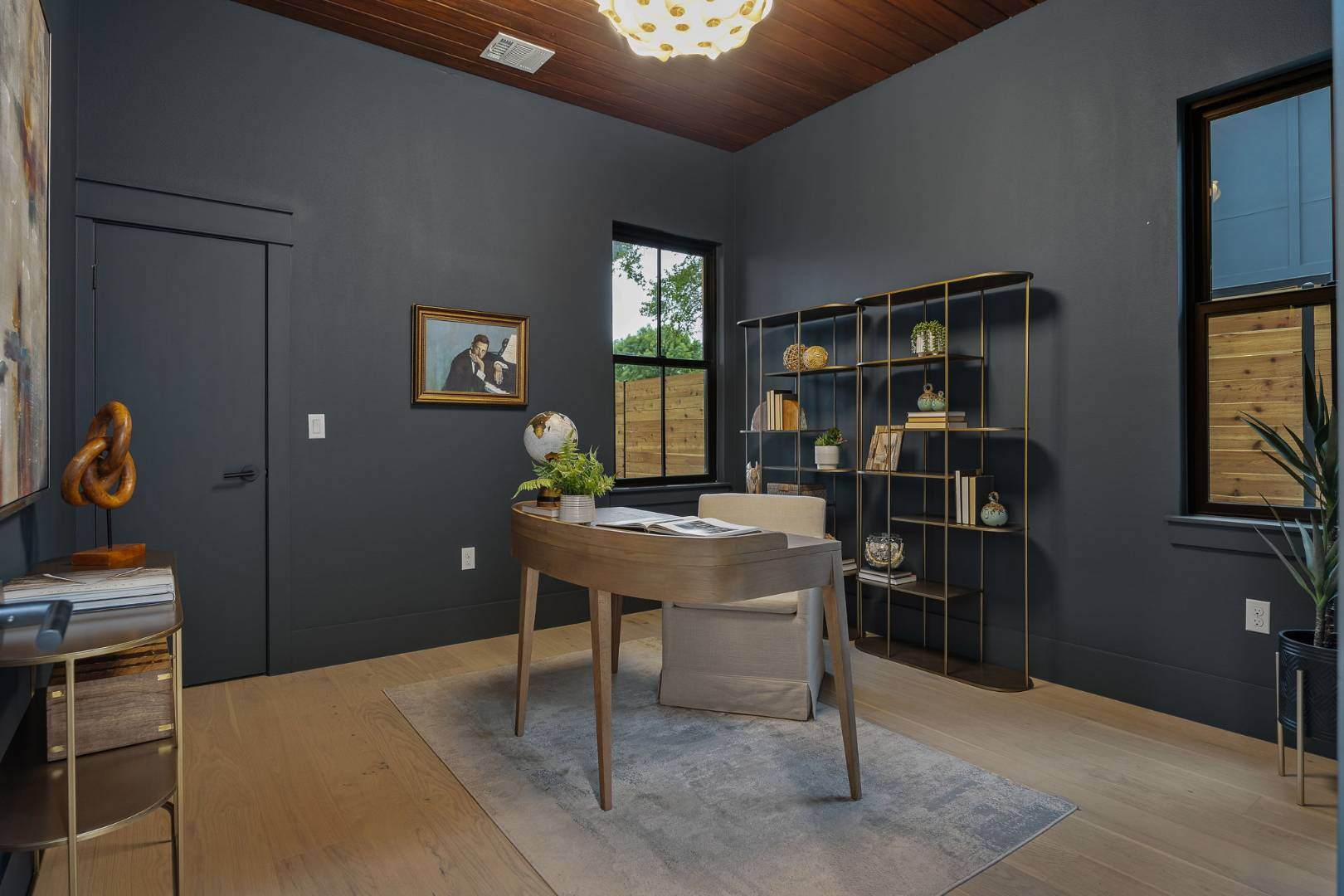 Sophisticated home office: Features a dark painted room with a wooden desk, gold shelving, and a variety of decor items, including a globe and framed artwork. | CKN Homes